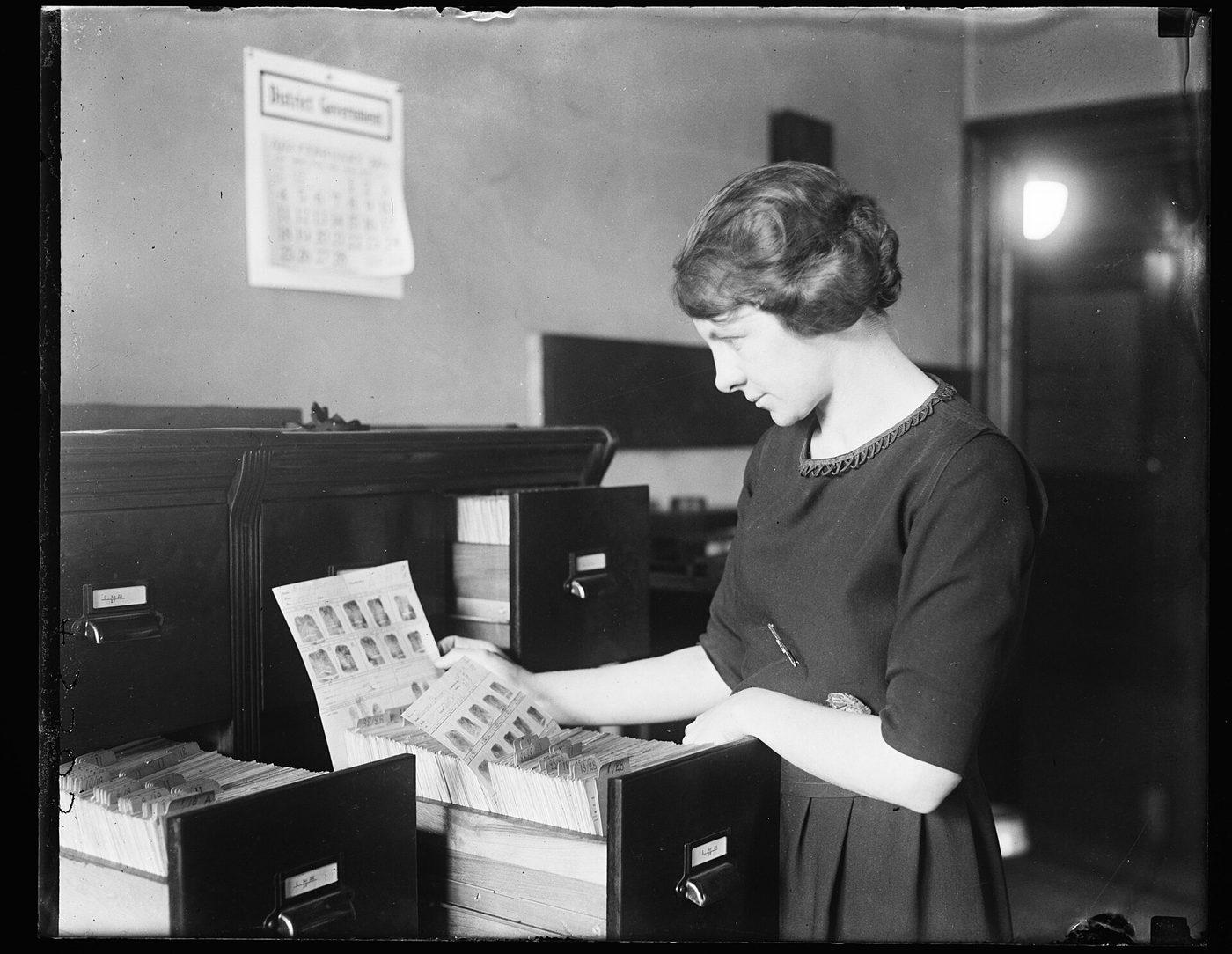 A young woman in a dark dress pulls a card of fingerprint and photo records from a drawer of a wooden card file cabinet