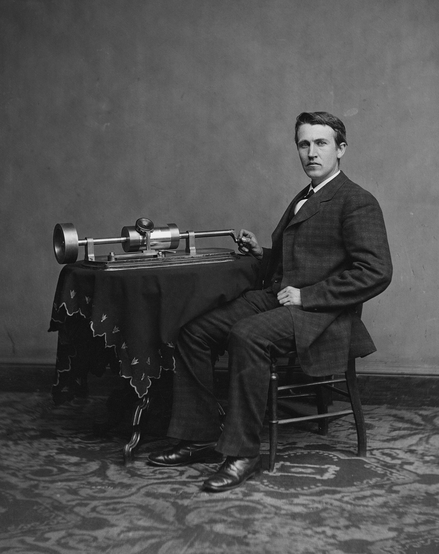 Thomas Edison sits beside a small table, his right hand on the crank of his early phonograph