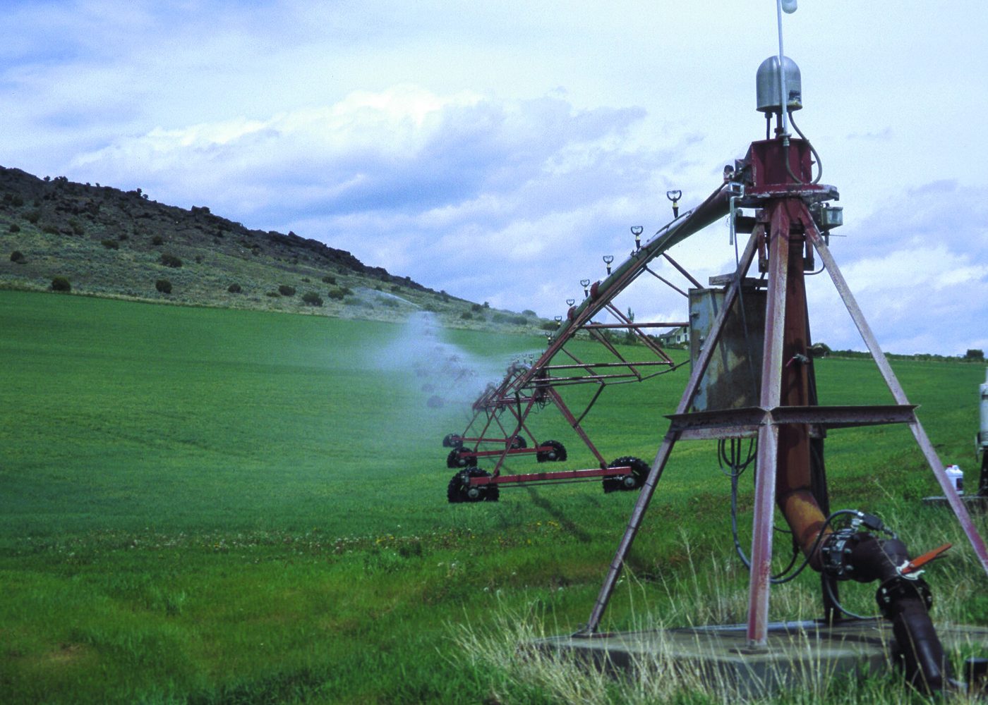 The red steel center tower of a pivot sprinkler, its arm spraying water across a green field below a sagebrush hill