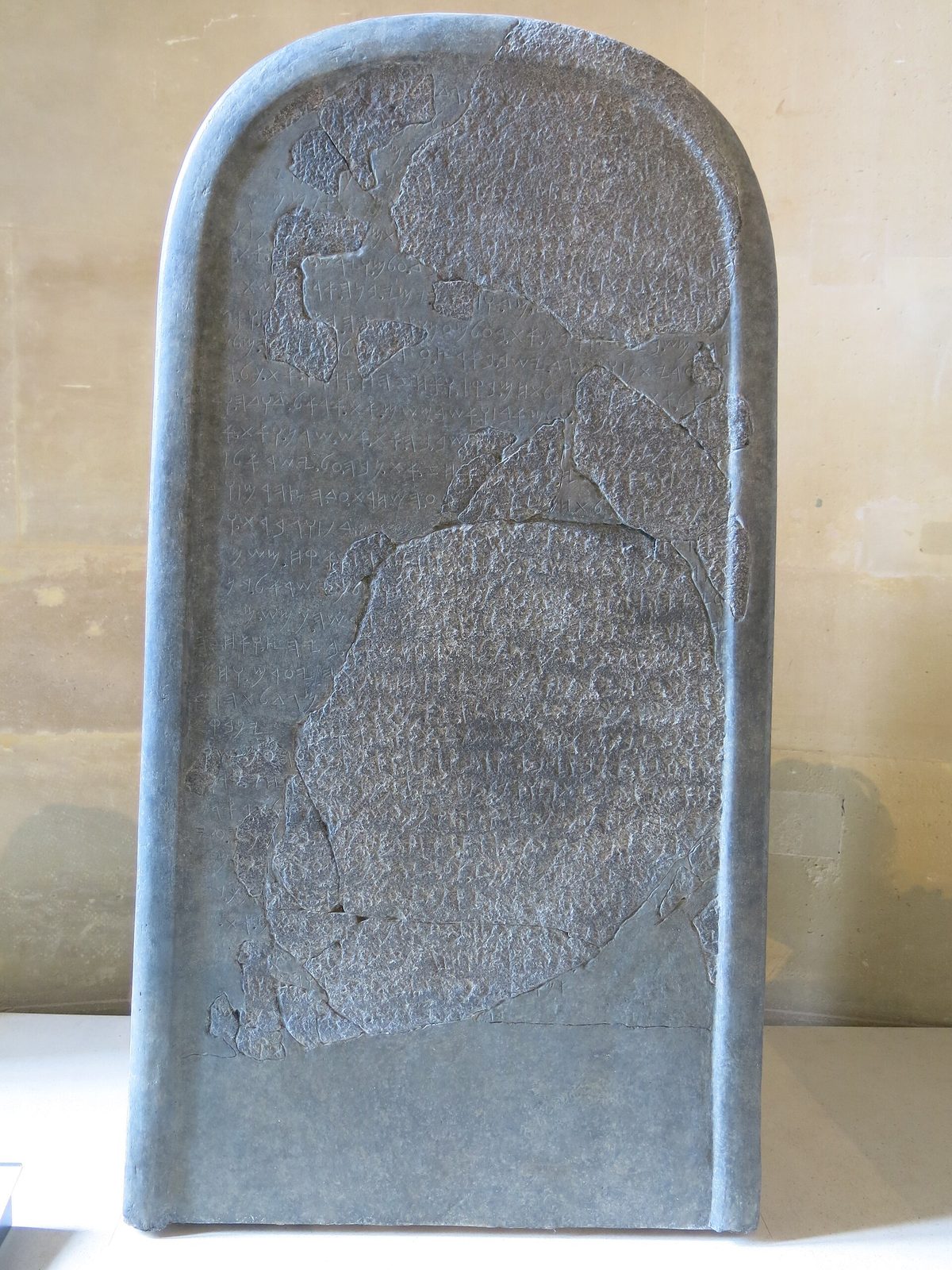A tall black basalt stone with a rounded top, covered in lines of old alphabetic writing, with some sections restored