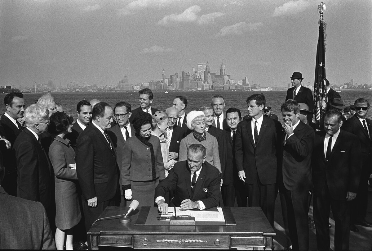 Black and white photograph of President Lyndon Johnson seated at a desk signing a bill on Liberty Island, surrounded by officials, with the Manhattan skyline behind them