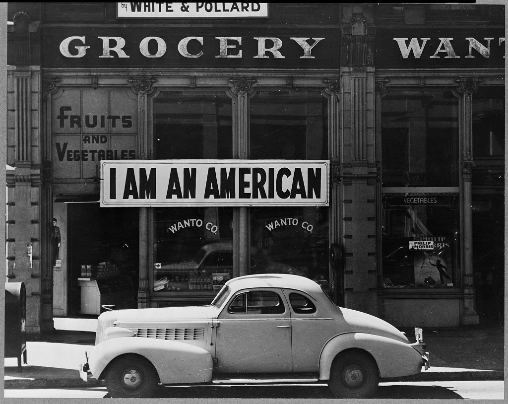 Black and white photograph of a closed grocery storefront in Oakland, California, with a large banner reading I AM AN AMERICAN above the window and a parked 1930s coupe in front