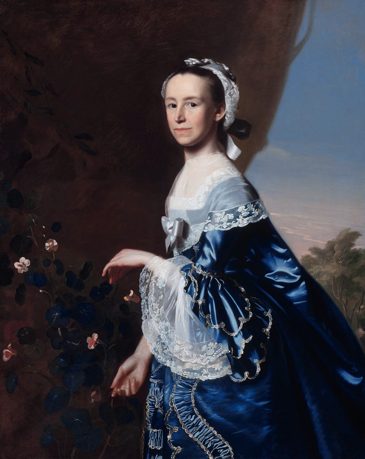Portrait of a woman in a blue satin dress with lace sleeves, standing and touching a flowering vine