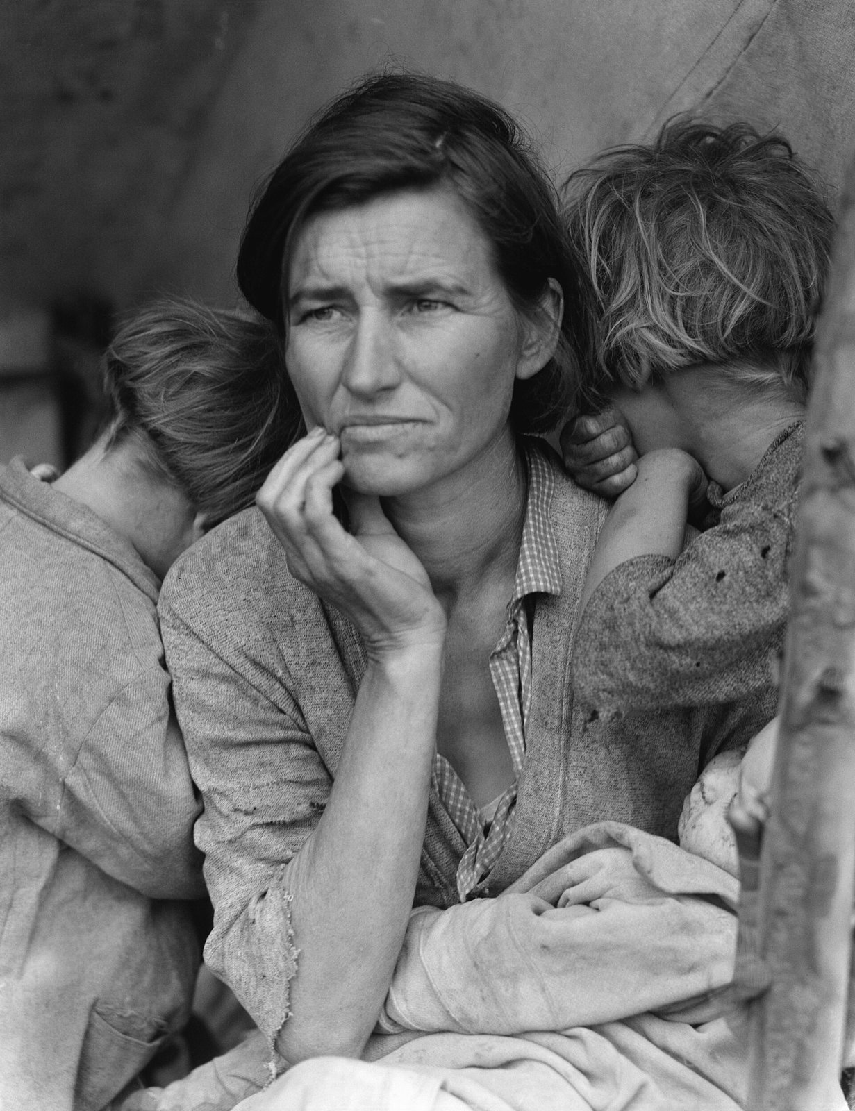 A worn mother in a migrant pea pickers' camp holds a baby while two children lean on her shoulders, their faces turned away