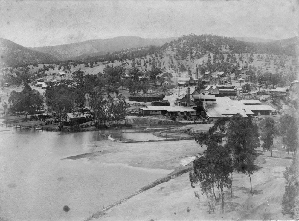 Old photograph of a tin smelter with smokestacks beside a dammed pond, with houses of a small mining town on the hills behind