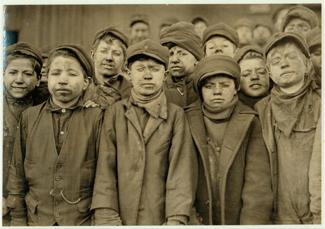 A crowd of boys in caps and work clothes, faces darkened with coal dust, packed together outside a coal breaker.