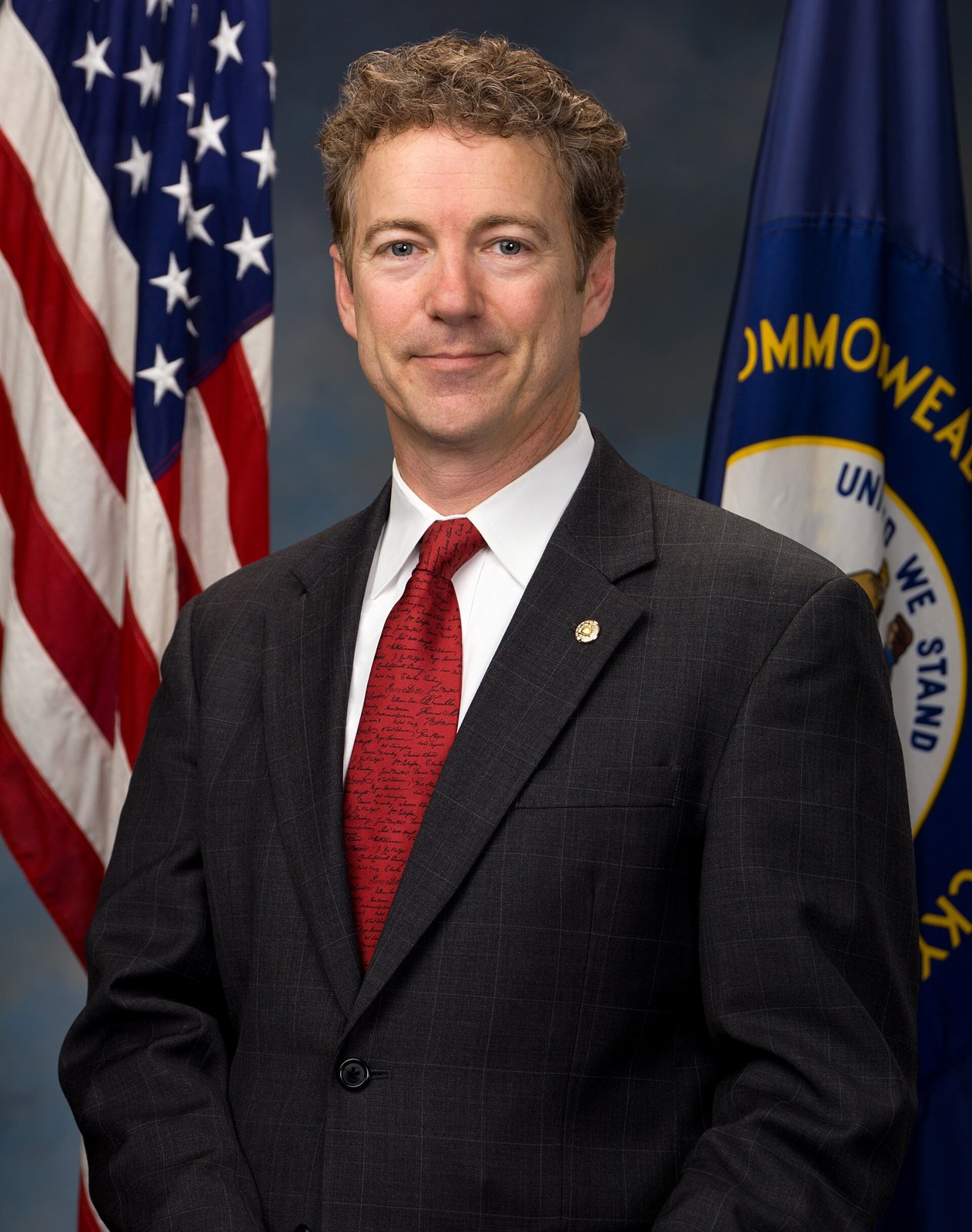 Official portrait of Senator Rand Paul in a dark suit and patterned tie, smiling, in front of a flag