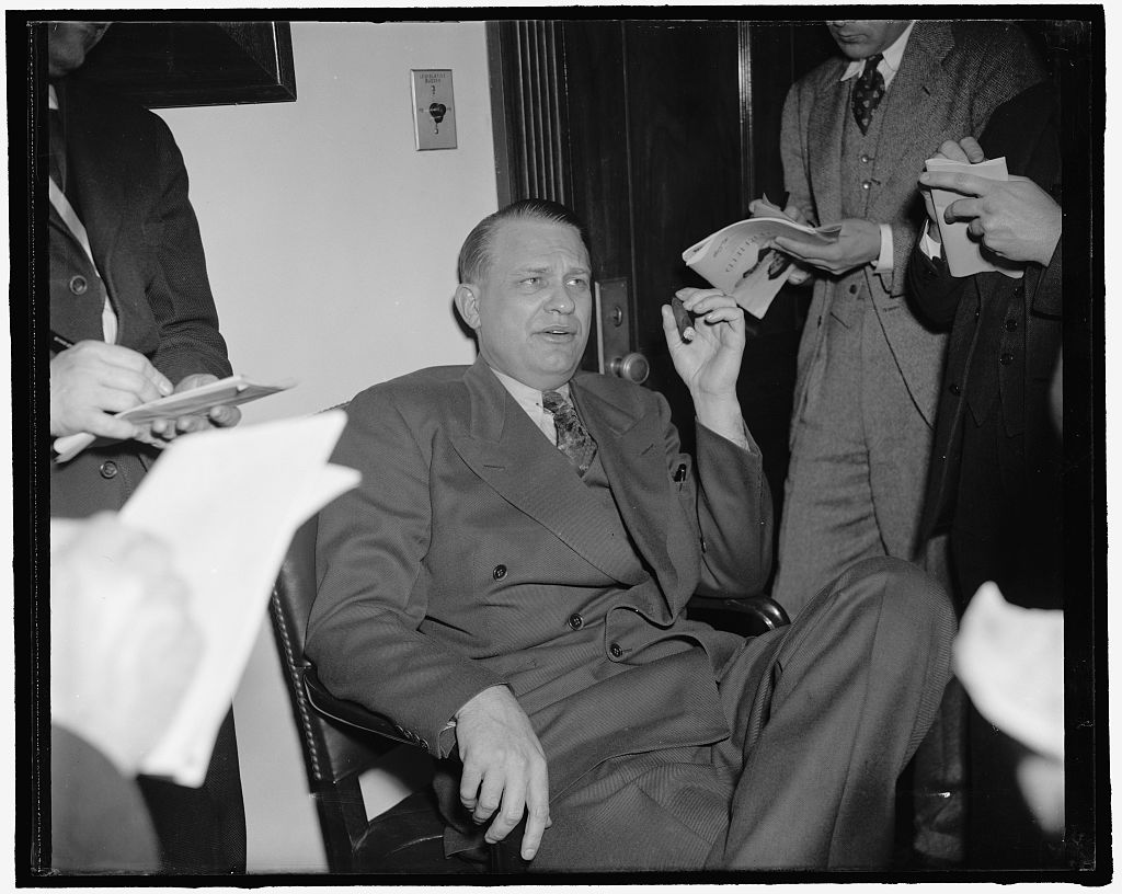 Black-and-white photograph of Martin Dies seated in a leather chair, holding a cigar and speaking, surrounded by standing men holding notepads