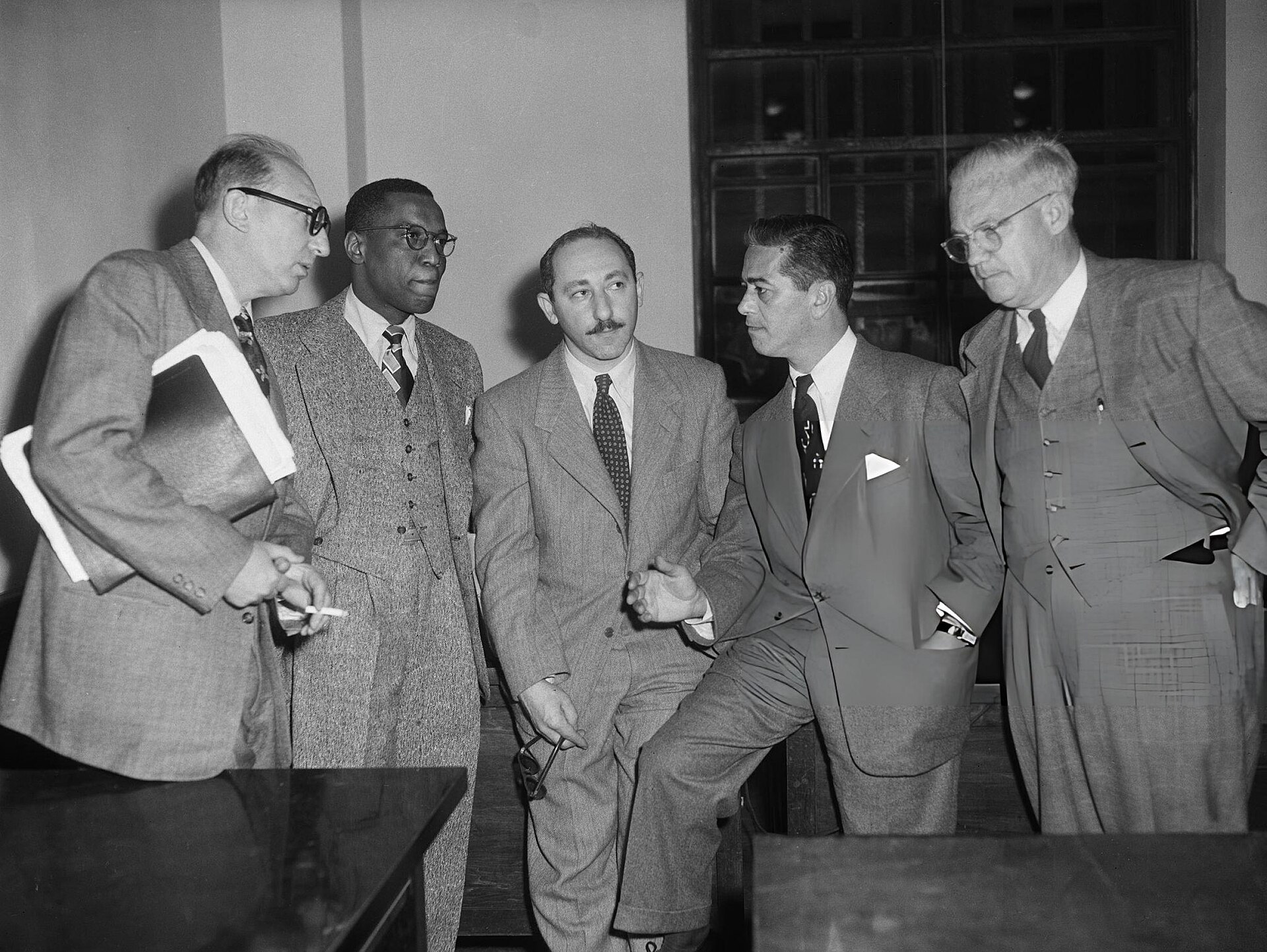 Press photograph of five men in suits and overcoats standing together on a city sidewalk