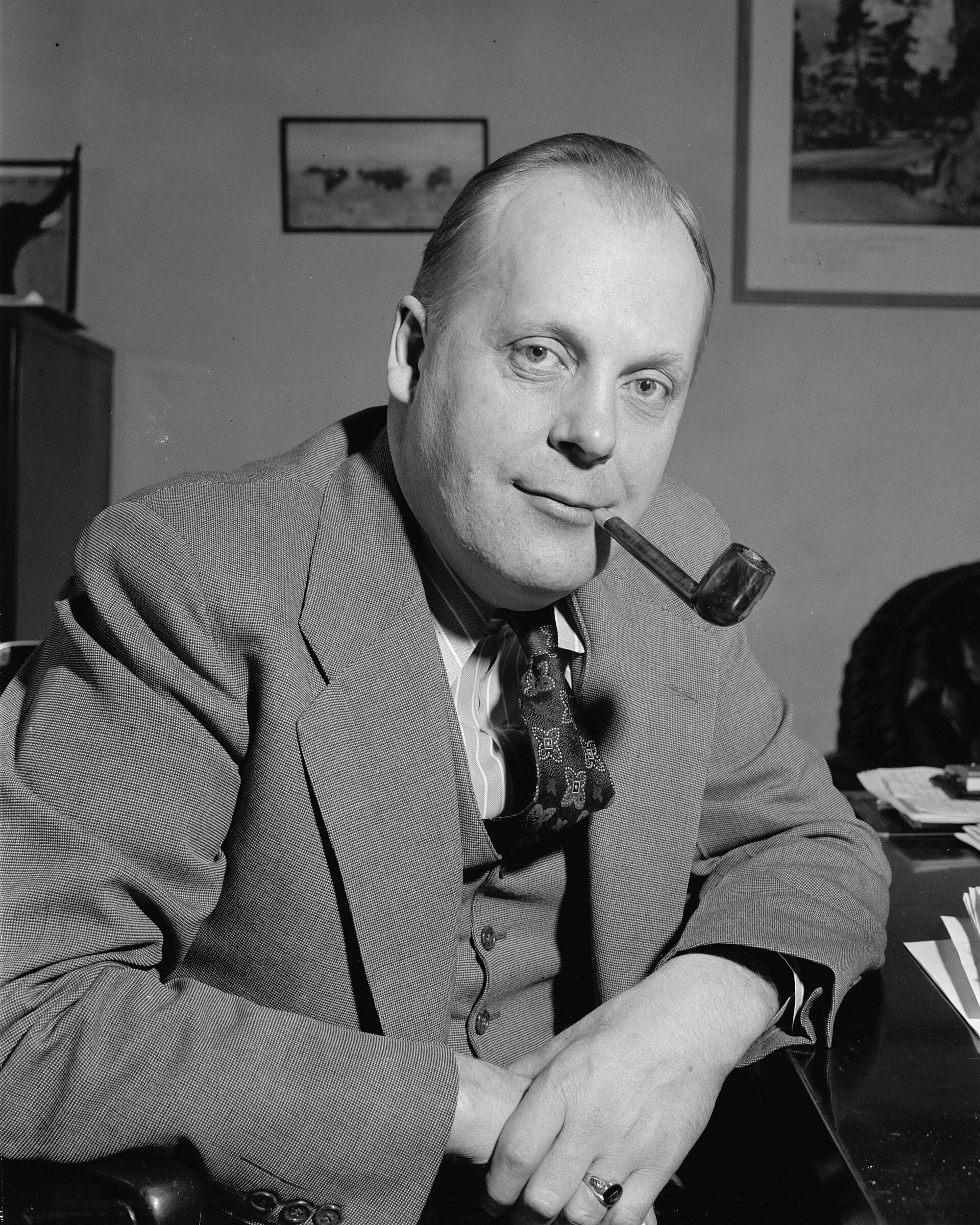 Black-and-white photograph of Karl Mundt seated at a desk in a light suit and patterned tie, smiling with a pipe in his mouth