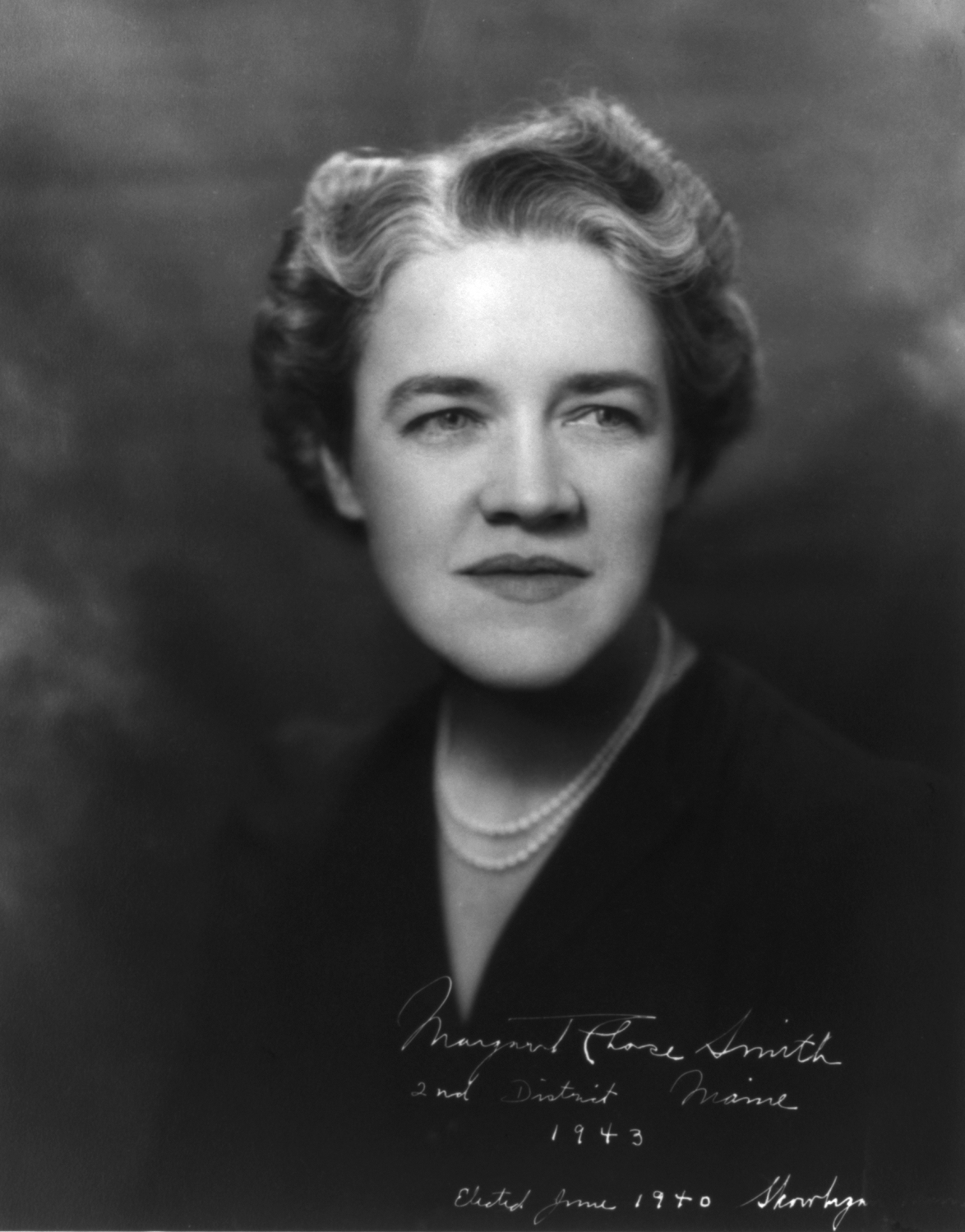 Black and white portrait of a woman with short wavy hair wearing a dark jacket, a white blouse with a ruffled collar and a corsage