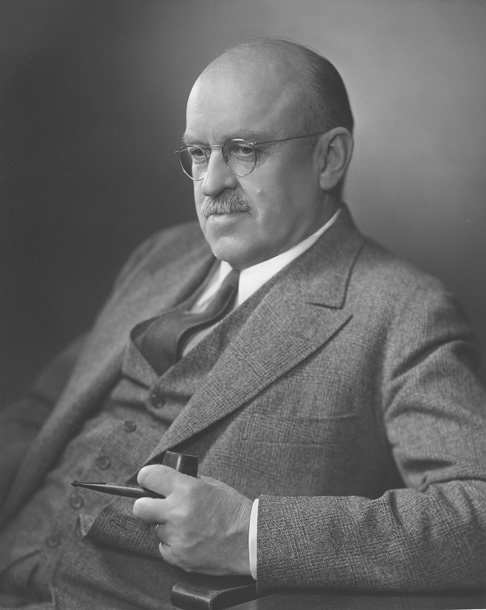 Head and shoulders portrait of an older man with white hair and round glasses, wearing a dark suit and tie