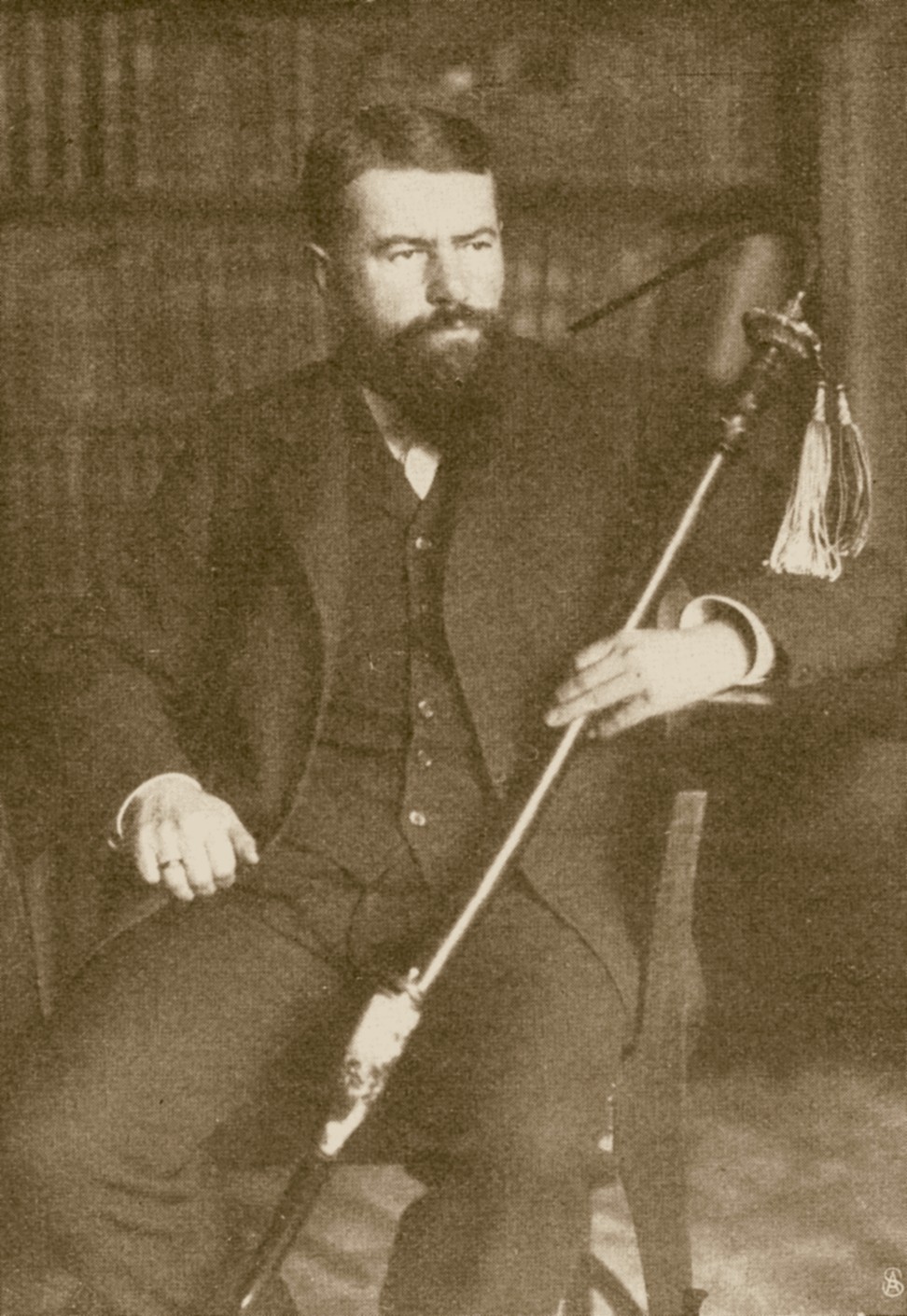 Sepia portrait photograph of Max Weber seated in a dark three-piece suit, with a full dark beard, holding a long tasseled staff across his lap, with bookshelves behind him
