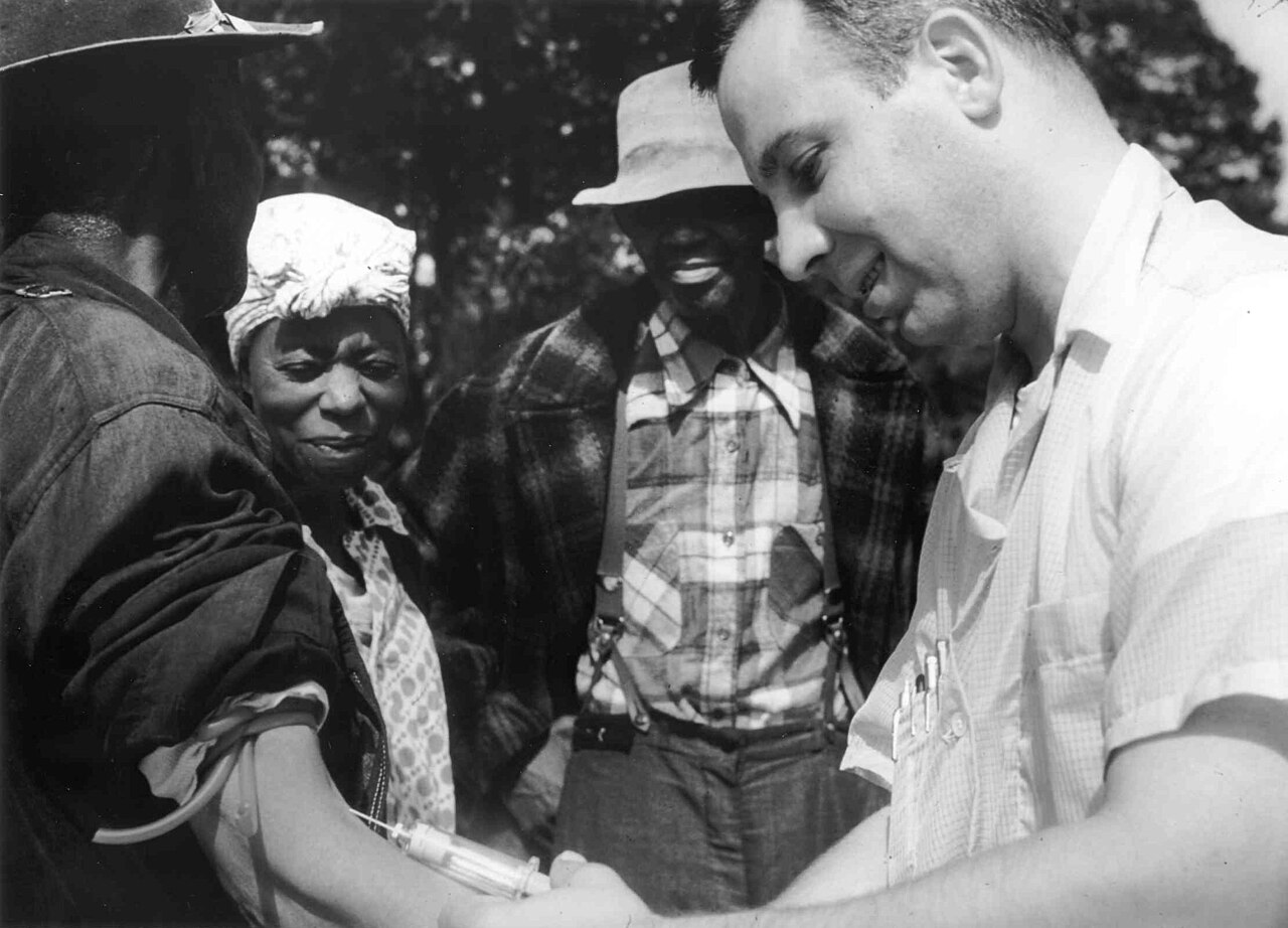 Black and white outdoor photograph of a white doctor in a short-sleeved shirt drawing blood with a syringe from the arm of a seated man, while a Black woman in a head wrap and a Black man in a hat and plaid shirt look on