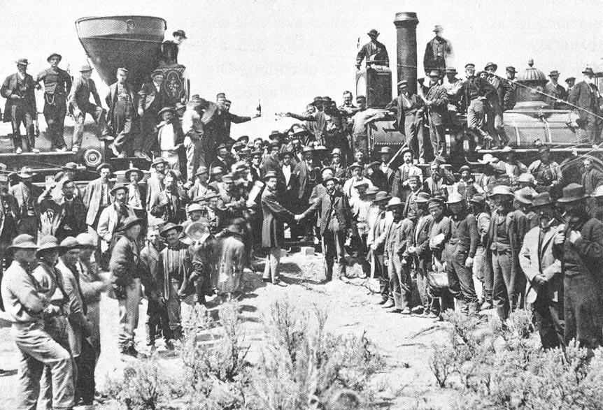 A crowd of workers and officials gathered around two locomotives meeting head to head on a single track, with men shaking hands and holding bottles aloft
