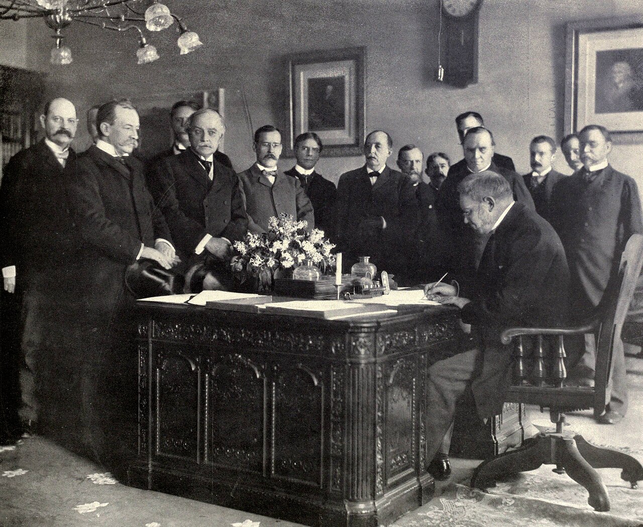 Printed page photograph of a bearded man seated at a desk signing a document while several men in formal dress stand around him