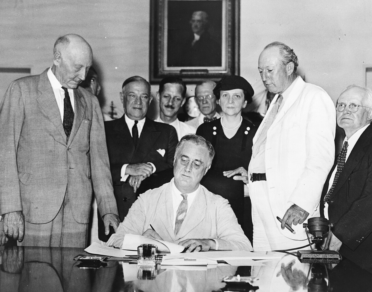 Black and white photograph of President Franklin Roosevelt seated at a desk signing a document while a group of men and one woman stand behind him