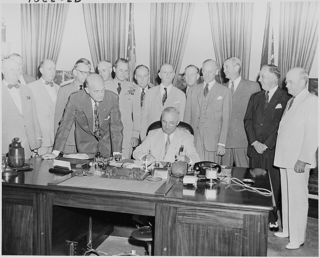 Black and white photograph of President Truman seated at his desk signing a bill while a group of men in suits and military uniforms stand behind him