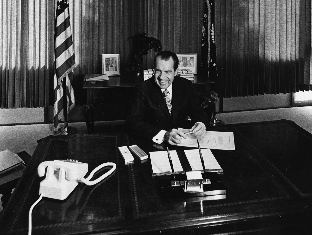 President Richard Nixon seated at a desk signing a document while several men in suits stand behind him