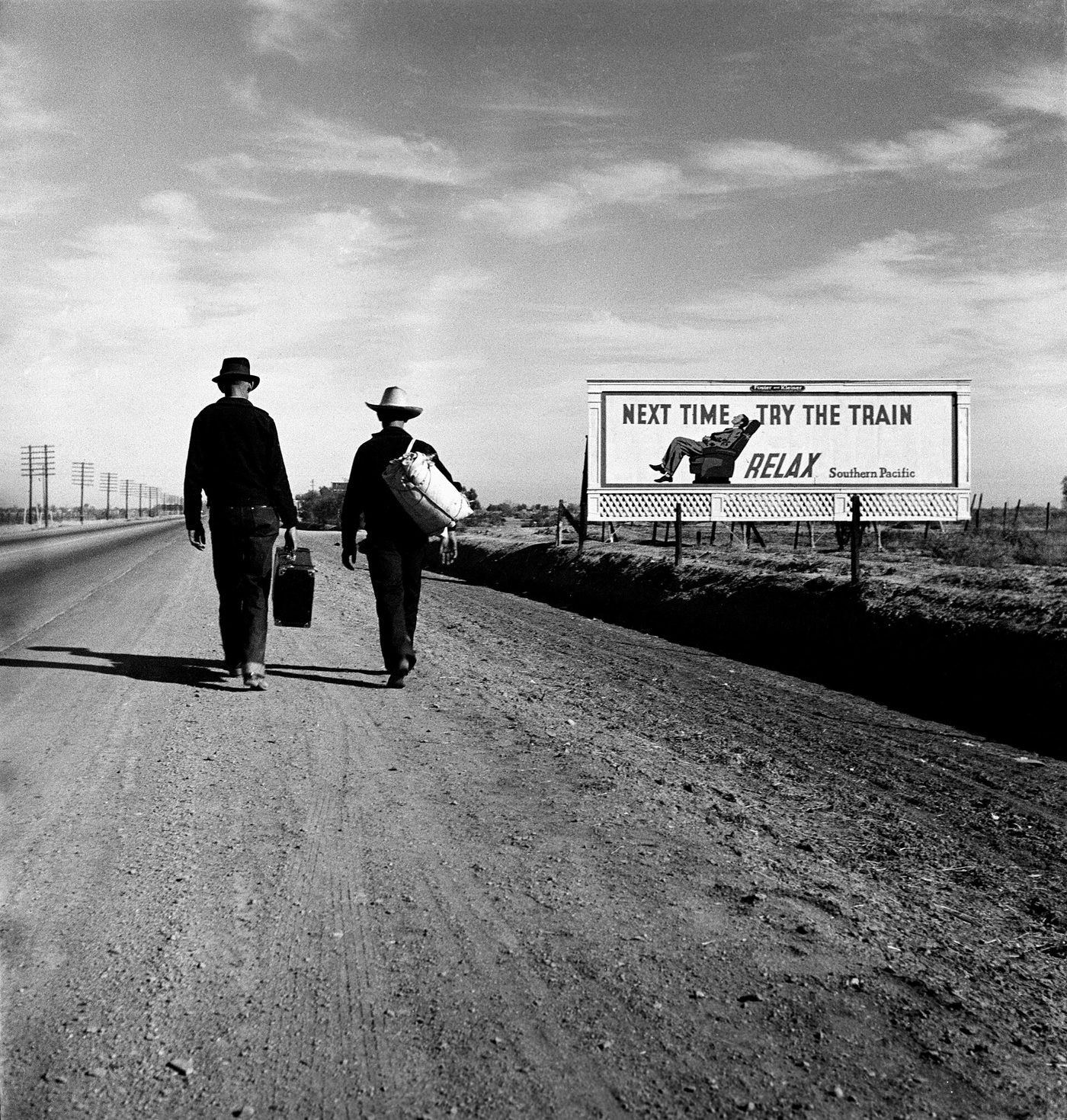 Two men walking along a highway past a large billboard reading Next time try the train, relax