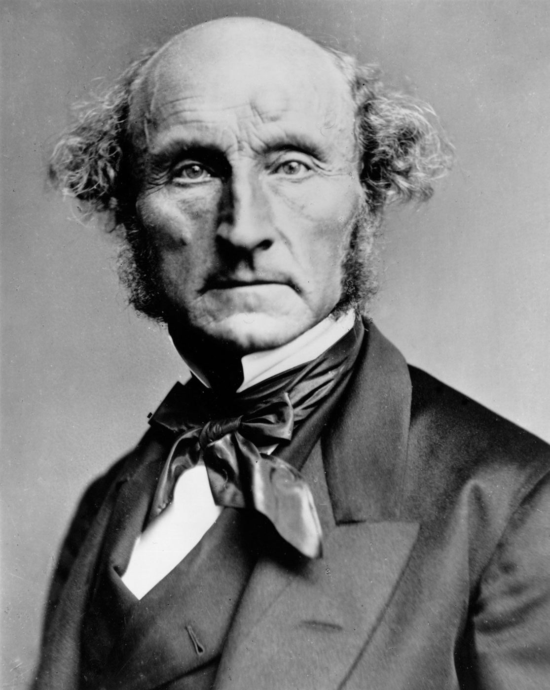 Photograph of John Stuart Mill in later life, bald, with side whiskers, wearing a dark coat